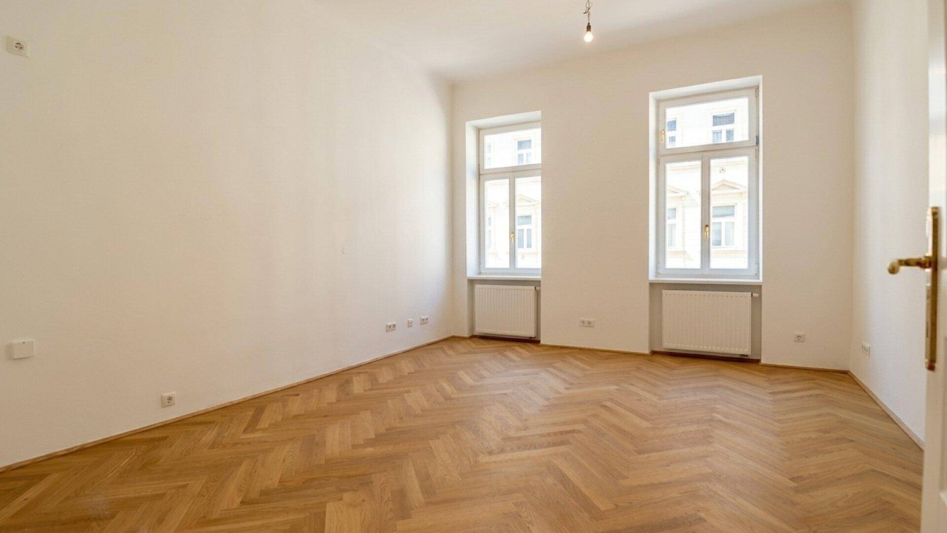 Geräumiges, helles Zimmer mit Fischgrätparkett, zwei Fenstern und einer weißen Tür.