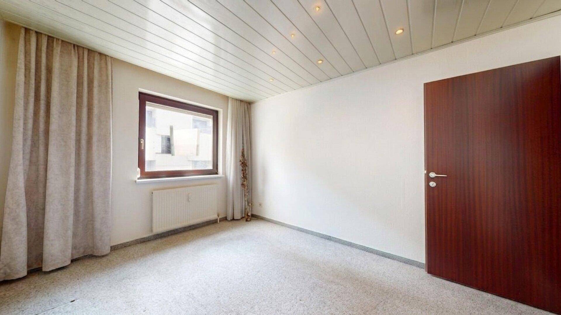 Schlafzimmer mit Teppichboden, Fenster und einer dunklen Holztür.