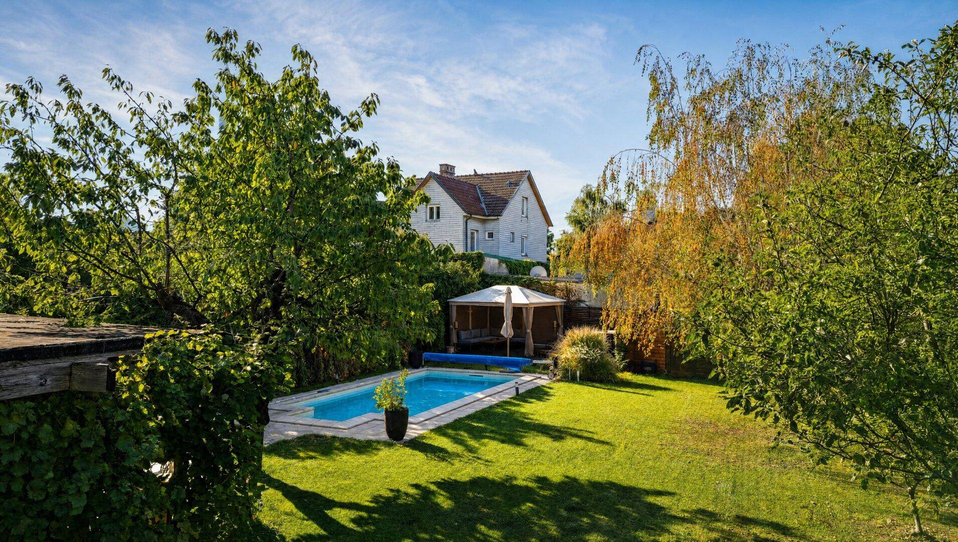 Gepflegter Garten mit Pool, umgeben von Bäumen und einem Haus im Hintergrund an einem sonnigen Tag.