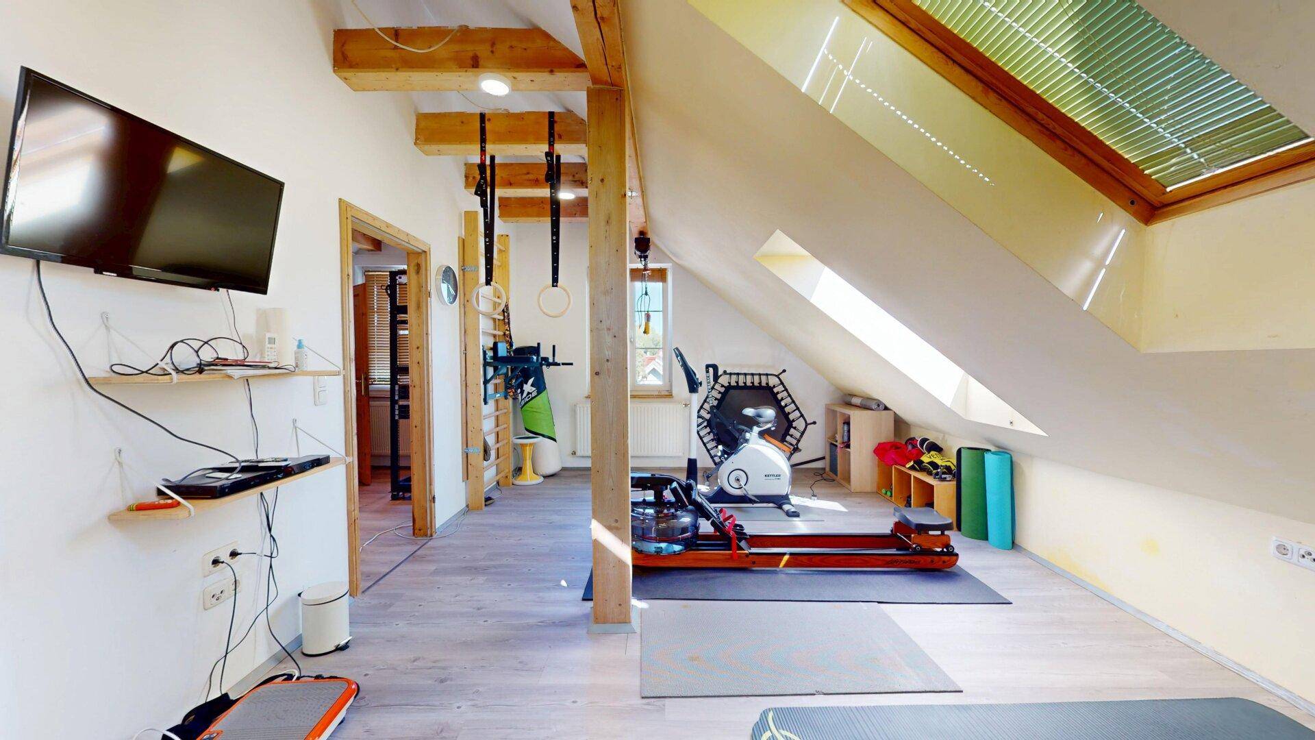 Heller Fitnessraum mit Holzbalken, Trainingsgeräten und einem Fernseher an der Wand.