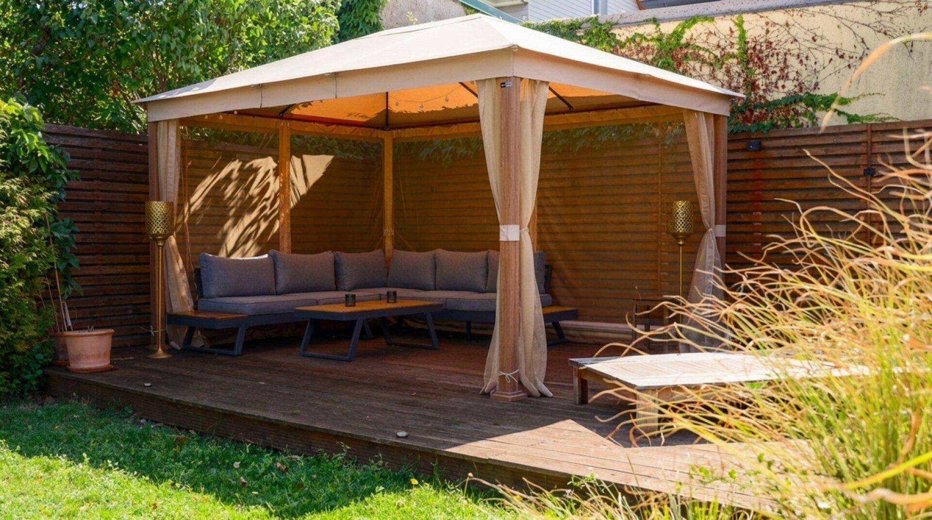 Geräumiger Pavillon mit Lounge-Möbeln auf einer Holzterrasse, umgeben von einem grünen Garten.