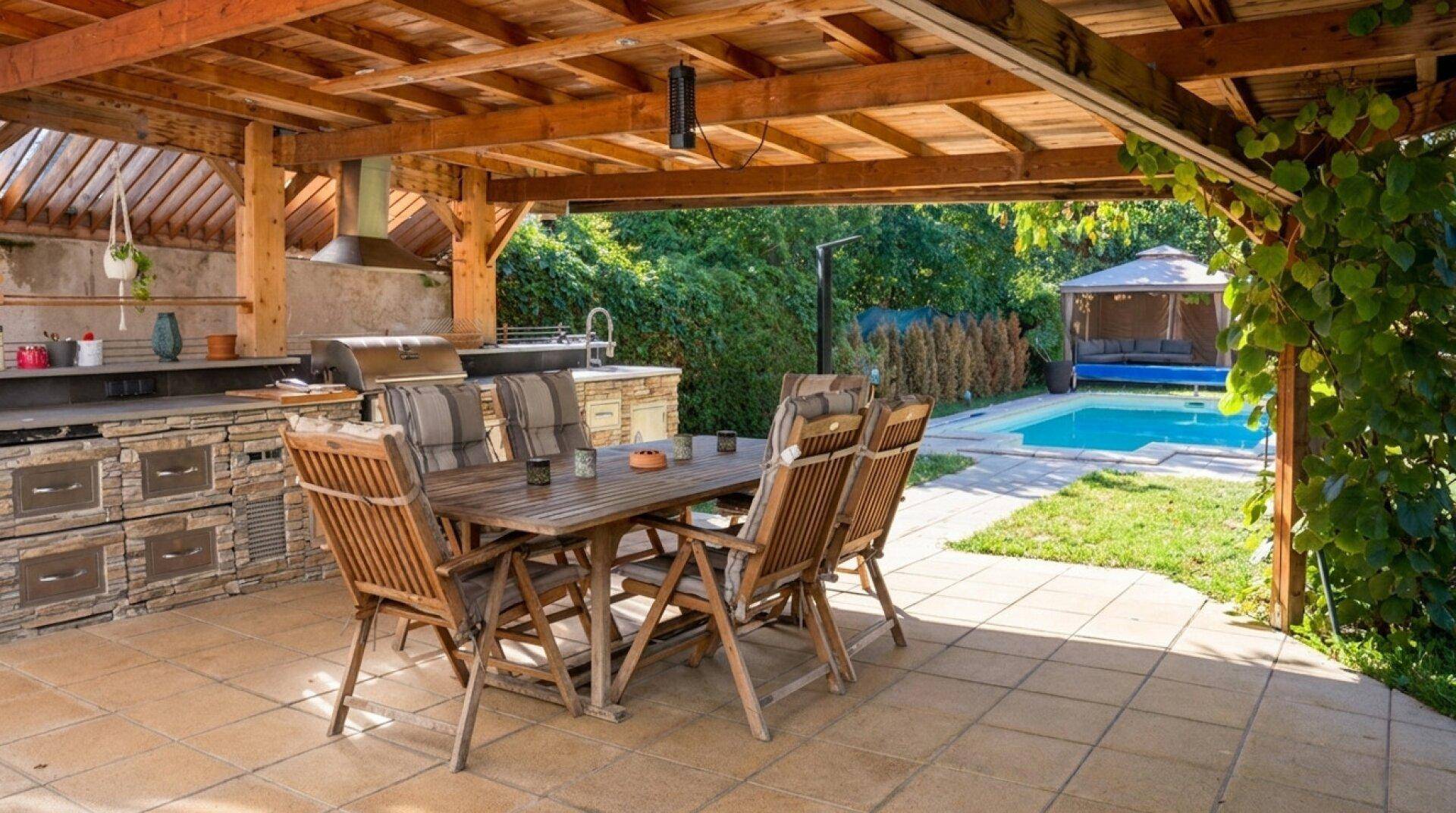 Gemütlicher Essbereich im Freien unter einer Holzpergola mit Blick auf den Pool und den Garten.