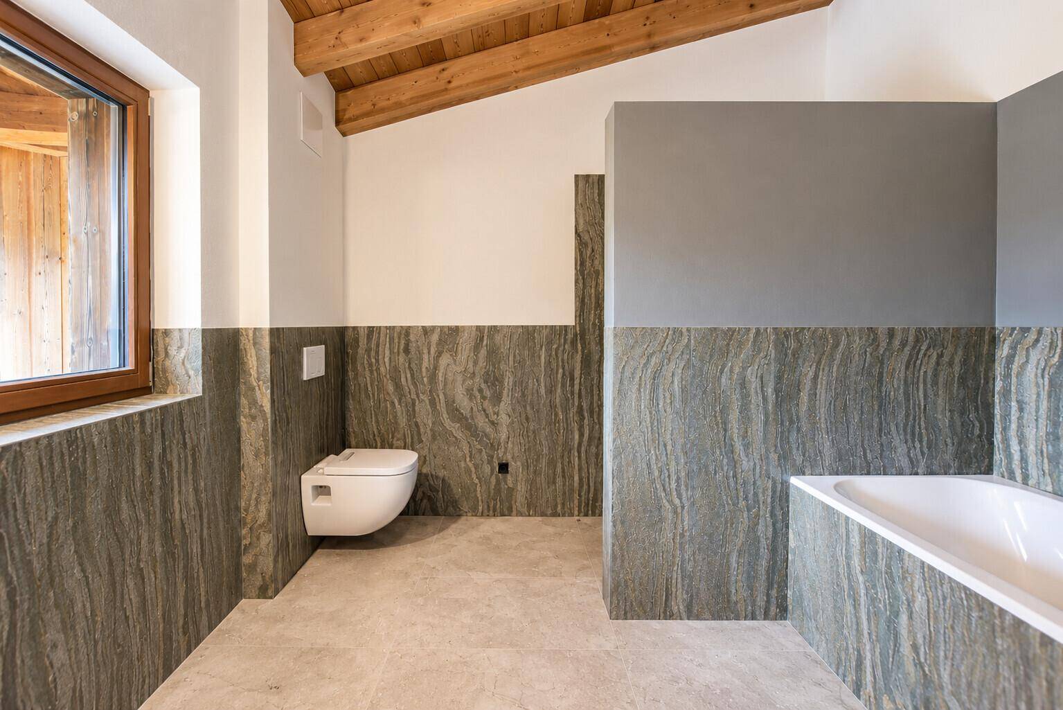 Modernes Badezimmer mit Natursteinfliesen, Badewanne und wandhängendem WC unter Dachschräge.