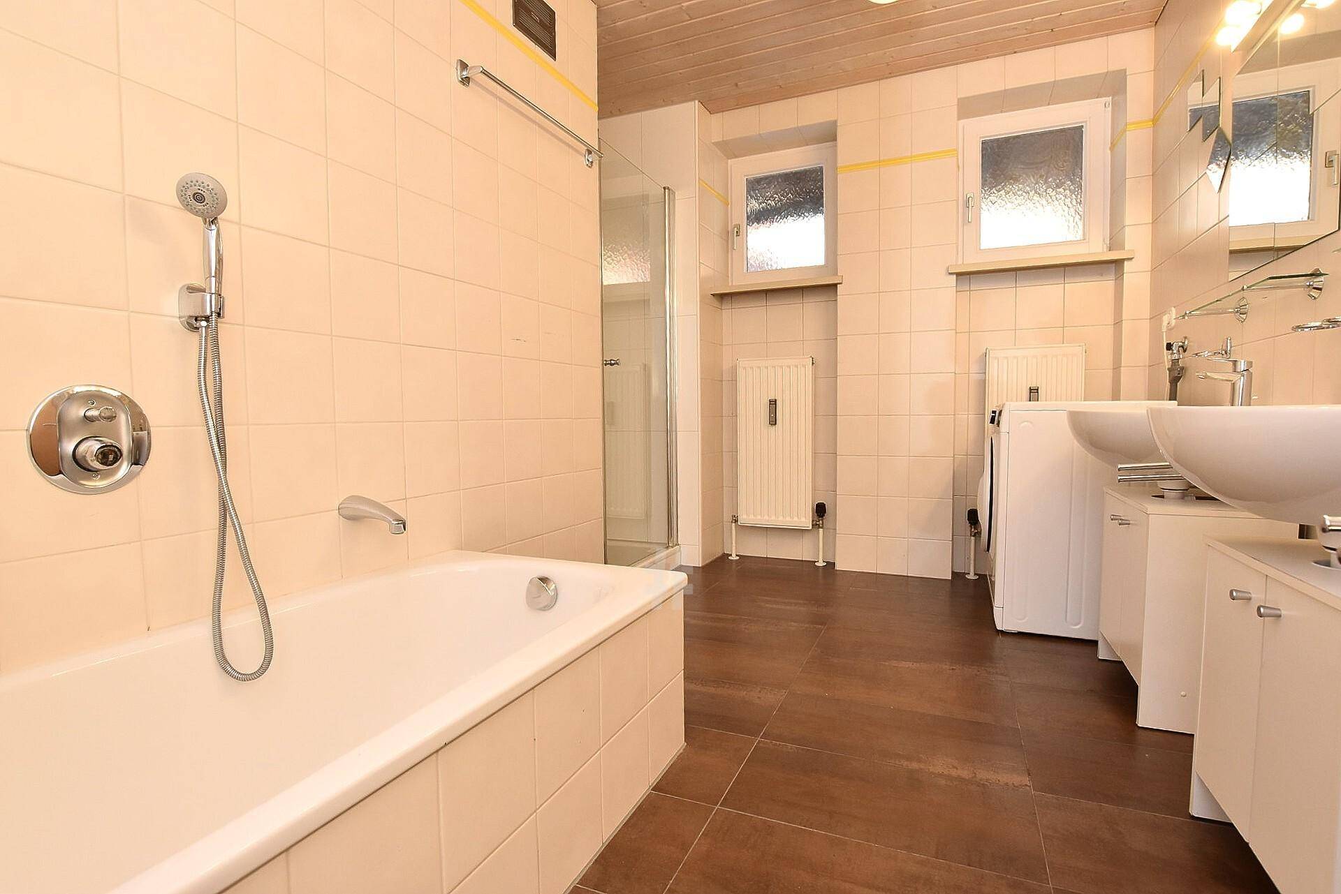Badezimmer mit Badewanne, separater Dusche, zwei Waschbecken und beigefarbenen Fliesen an den Wänden.