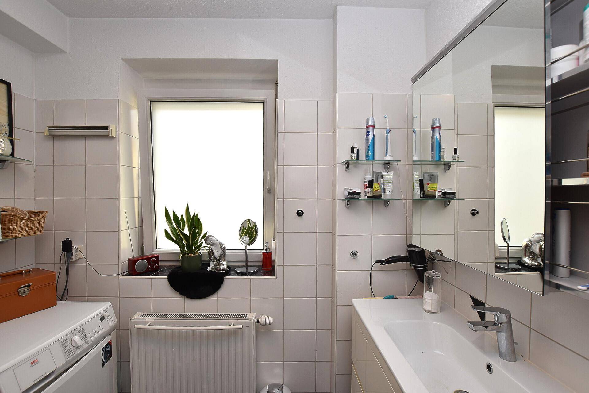 Funktionales Badezimmer mit weißen Fliesen, Waschbecken, Spiegelschrank und Fenster.