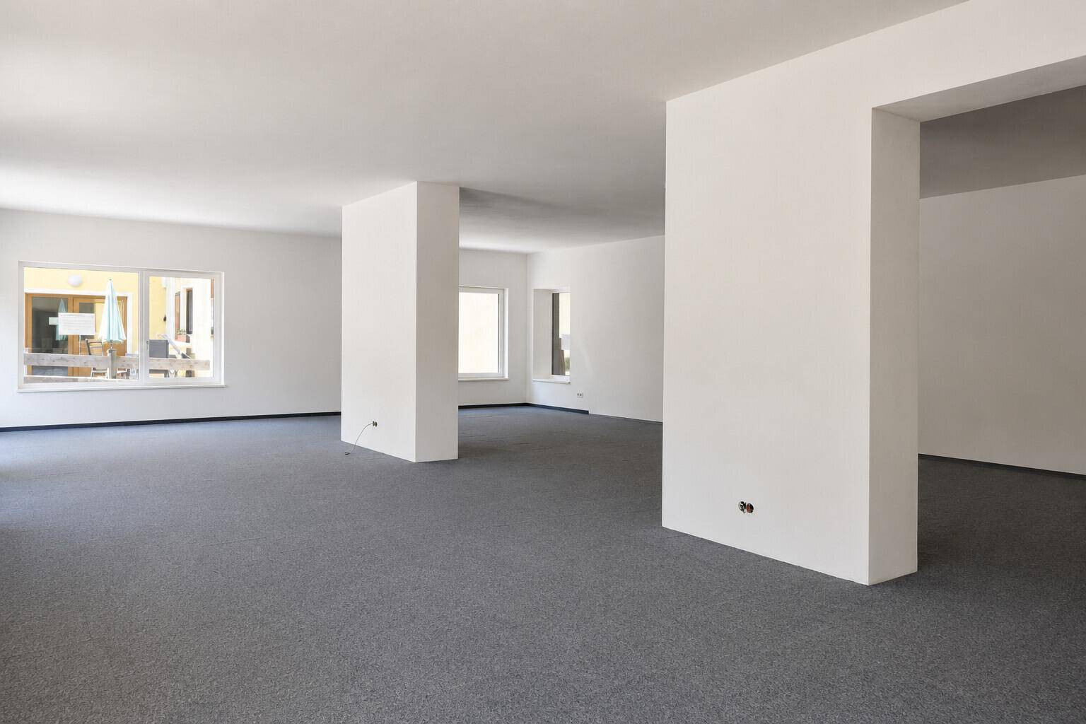 Heller, offener Gewerberaum mit weißen Wänden und grauem Teppichboden, ideal für Büros oder Ausstellungsflächen.