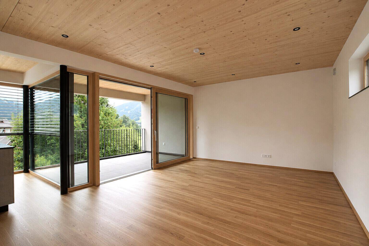 Heller, leerer Wohnraum mit Holzboden, Holzdecke und Schiebetüren zum Balkon.