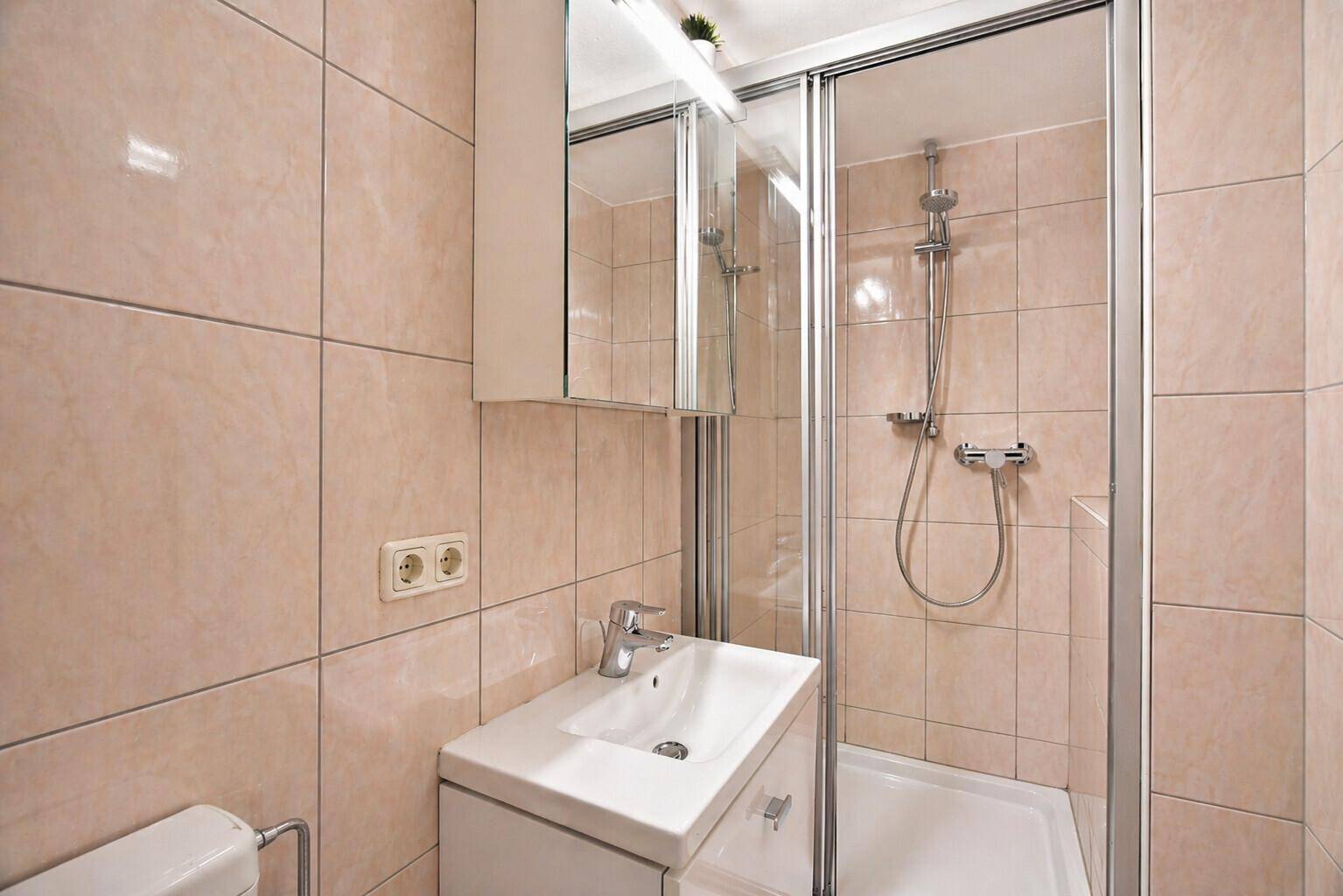Praktisches Badezimmer mit beigen Fliesen, Duschkabine, Waschtisch und Spiegelschrank.