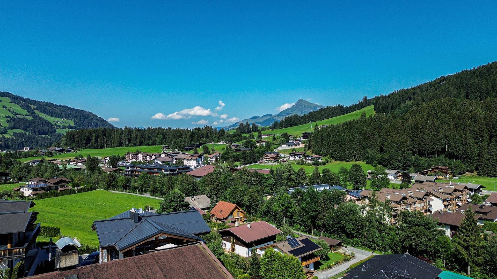 Panoramablick über eine idyllische Siedlung in einem grünen Tal mit bewaldeten Hügeln und Bergen im Hintergrund.