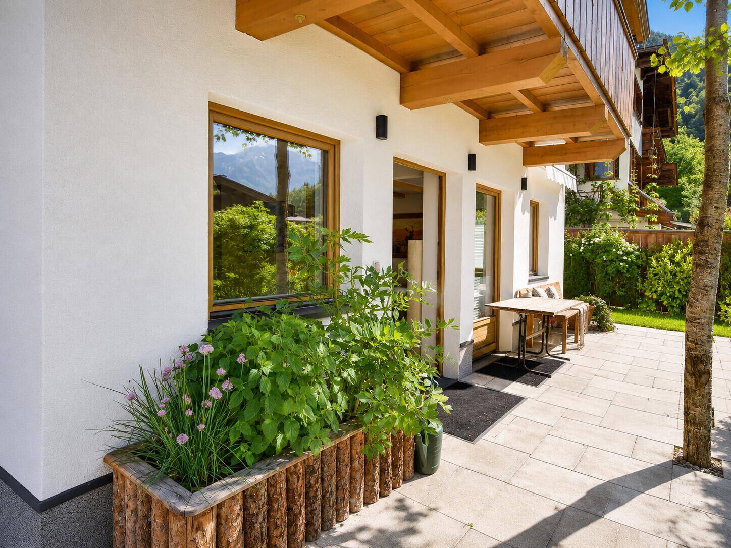 Sonniger Terrassenbereich am Haus mit Sitzgelegenheiten und Blumenbeeten.
