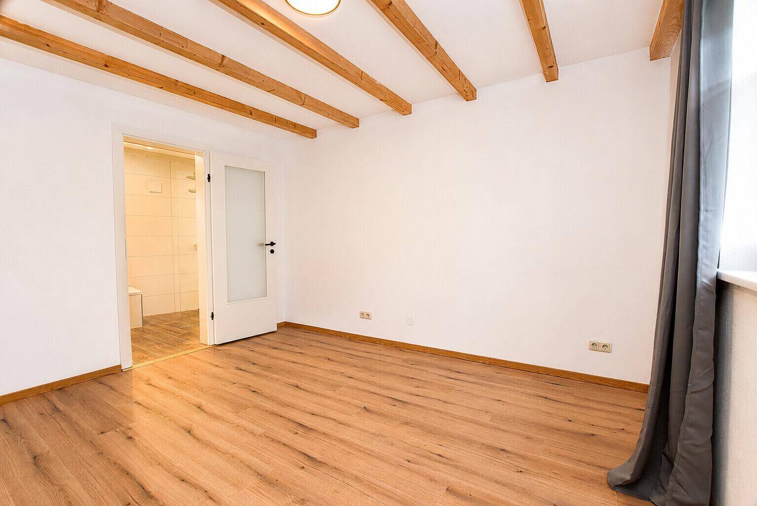 Schlafzimmer mit Holzbalken, Holzboden und Zugang zum angrenzenden Badezimmer.