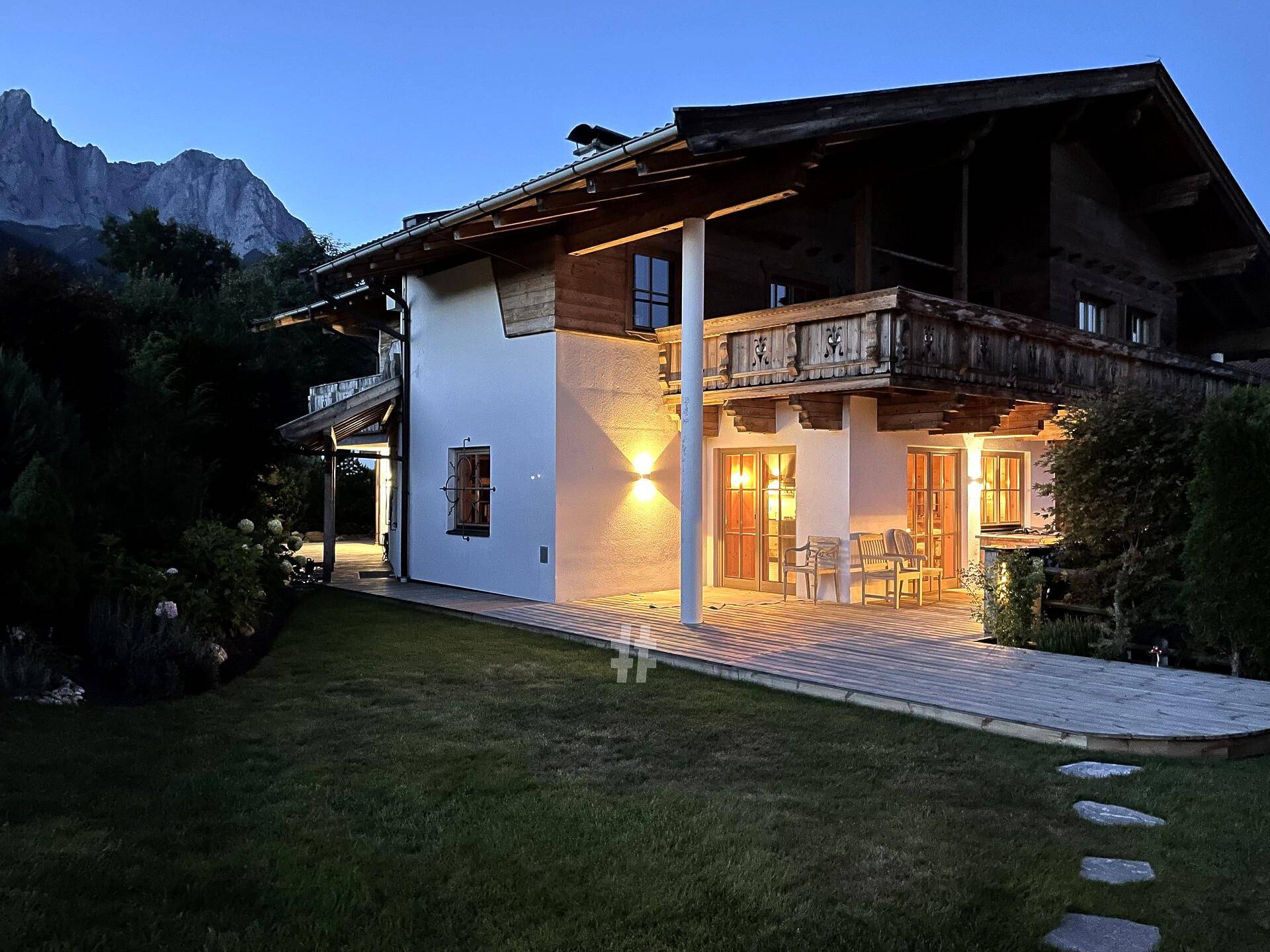 Beleuchtetes Chalet mit großer Holzterrasse und gepflegtem Garten in der Abenddämmerung.