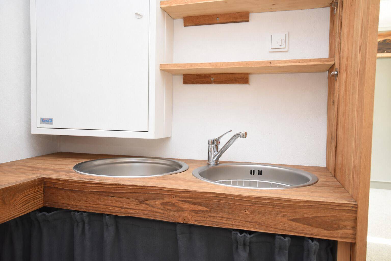 Kleine Küchenzeile mit zwei Edelstahlspülbecken, einer Holztheke und einem weißen Hängeschrank.