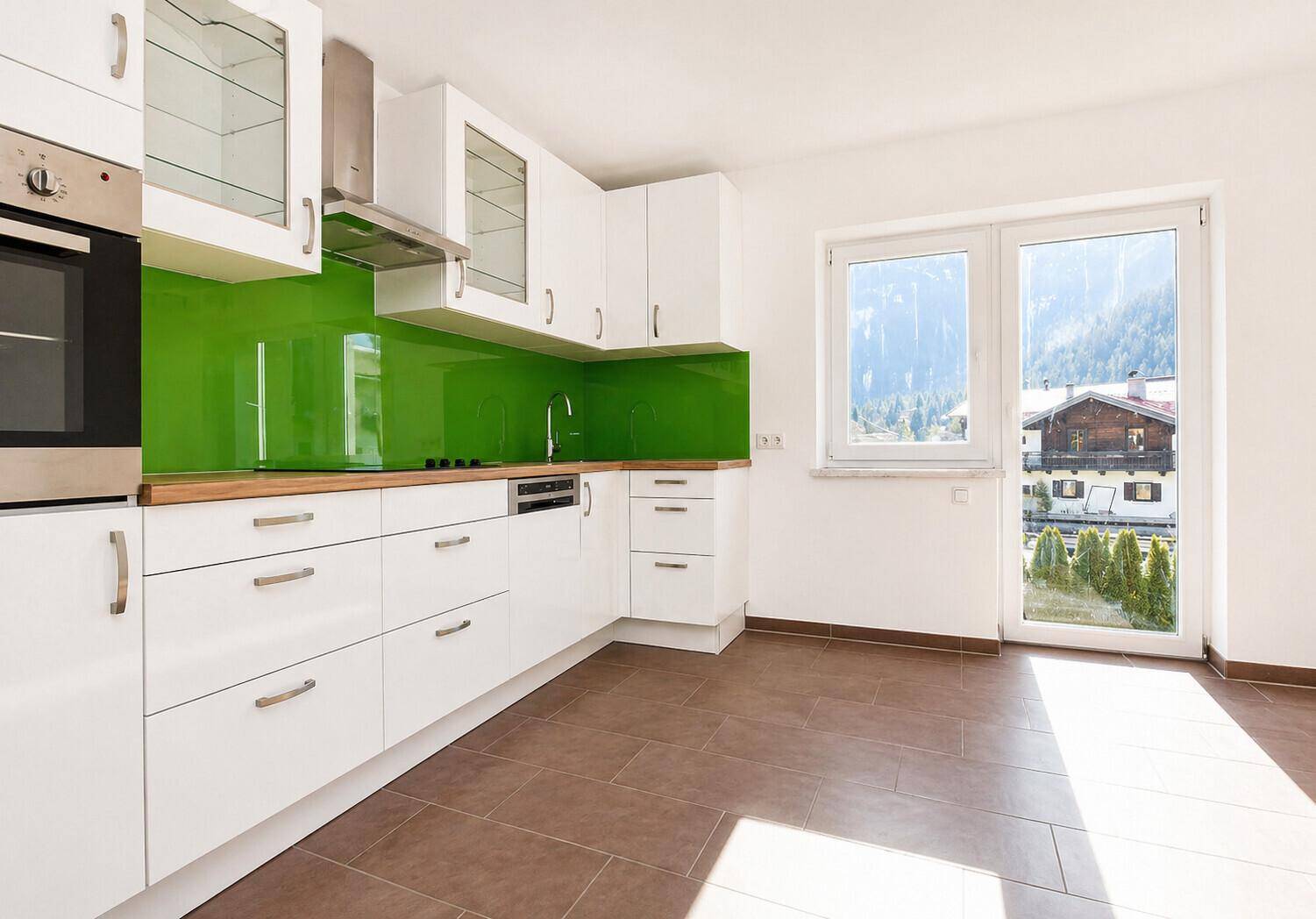 Helle Küche mit Bergblick, weißen Schränken, grüner Spritzschutzwand und Fenster.