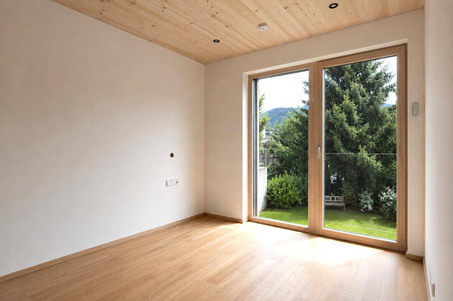 Ruhiges Zimmer mit Holzboden, Holzdecke und großem Fenster zum grünen Gartenbereich.