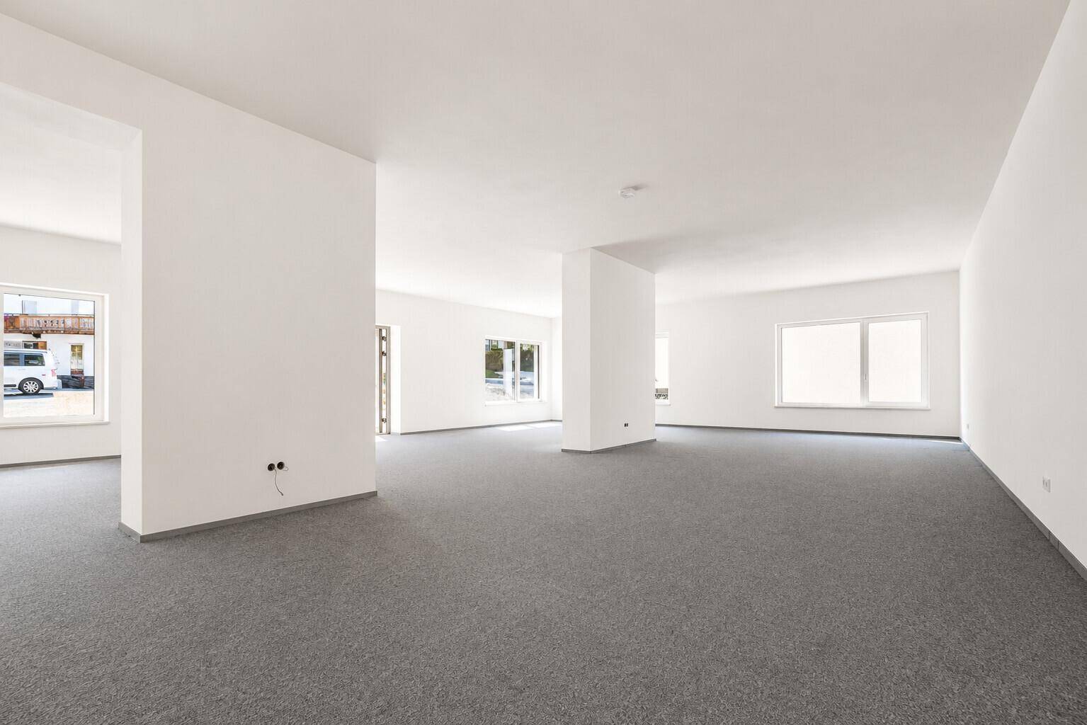 Geräumiger, leerer Gewerberaum mit weißen Wänden und grauem Teppichboden, ideal für Büros oder Praxen.
