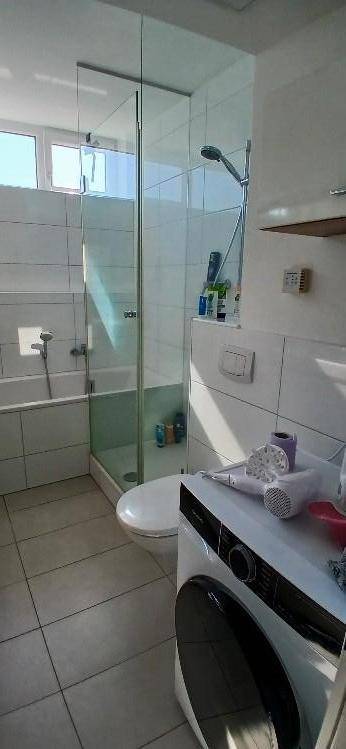 Badezimmer mit Duschkabine, Badewanne und integrierter Waschmaschine, hell durch Fenster.