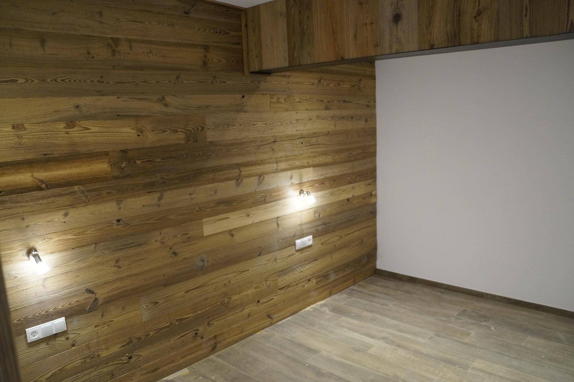 Raum mit einer markanten Holzwandverkleidung, weißen Wänden und modernem Fliesenboden, beleuchtet von Wandlampen.