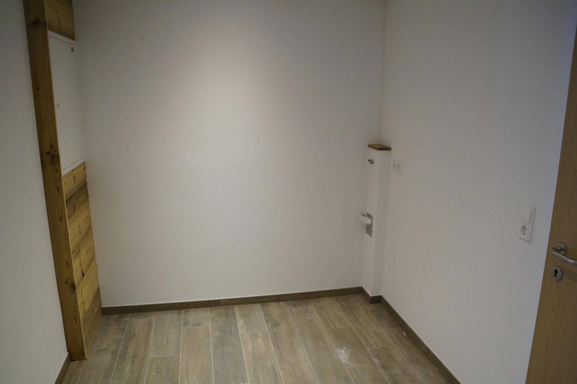 Leerer Raum mit weißen Wänden, Holzfliesenboden und einer Wasseranschlussmöglichkeit an der Wand.