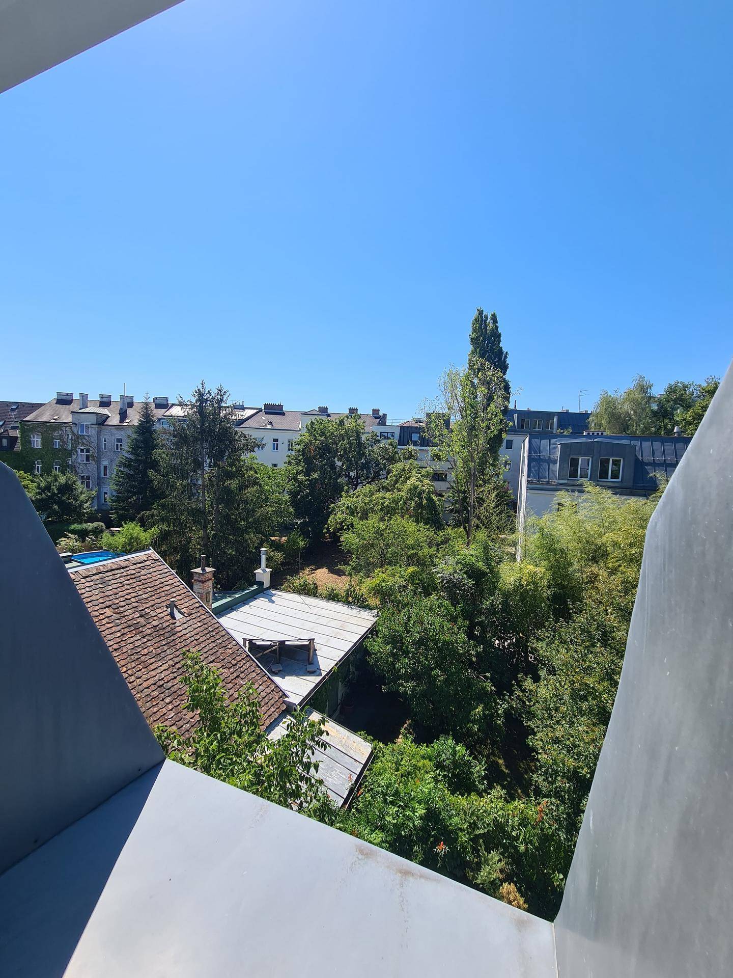 Ausblick von einem Balkon auf umliegende Gebäude und grüne Bäume.
