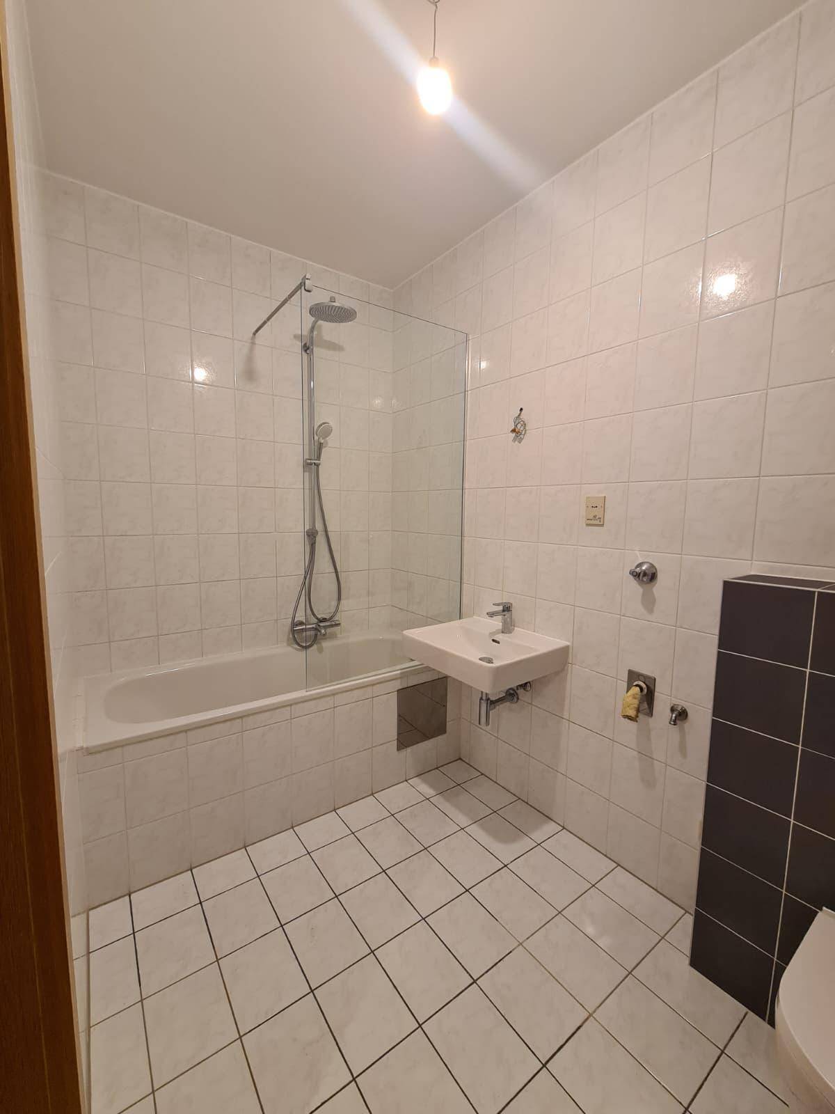 Badezimmer mit weißen Fliesen, Badewanne mit Duschfunktion und kleinem Waschbecken.