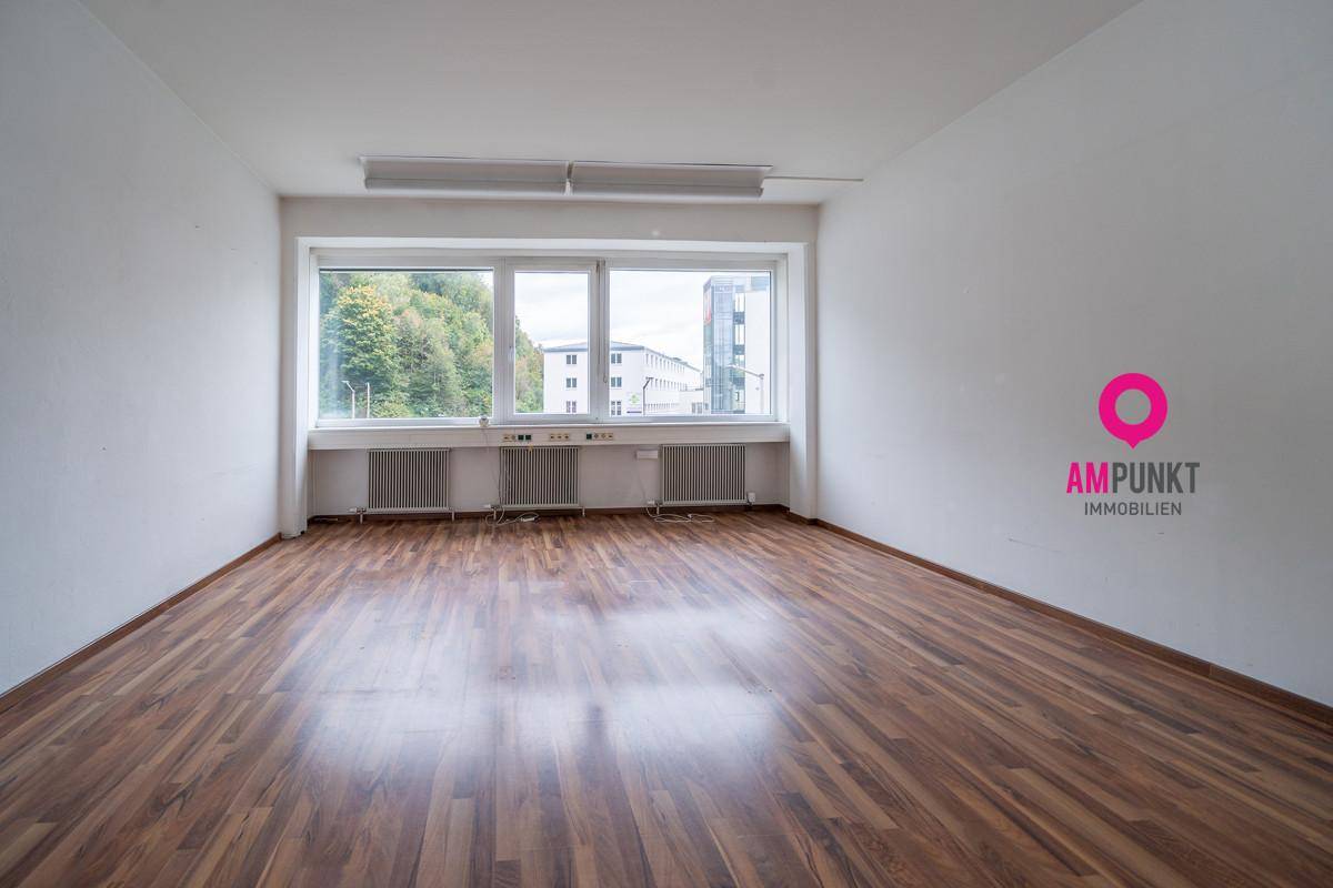 Heller Büroraum mit Holzboden und großen Fenstern, die einen Blick auf Bäume und Gebäude bieten.