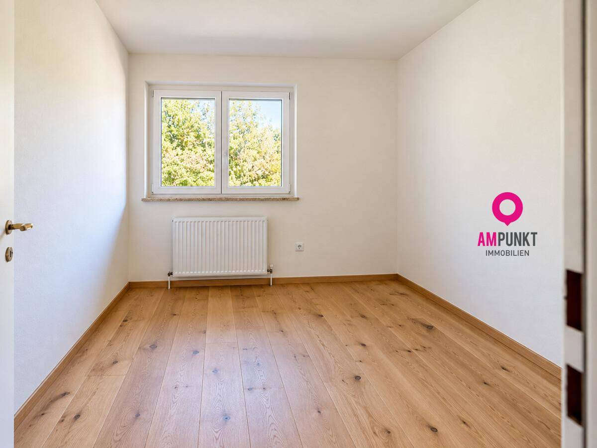 Freundliches Zimmer mit Holzboden, weißen Wänden und Fenster mit Blick auf grüne Bäume.
