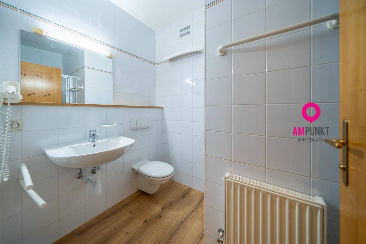 Helles Badezimmer mit weißen Fliesen, Waschbecken, WC und Heizkörper, ausgestattet mit Haltegriffen für Barrierefreiheit.