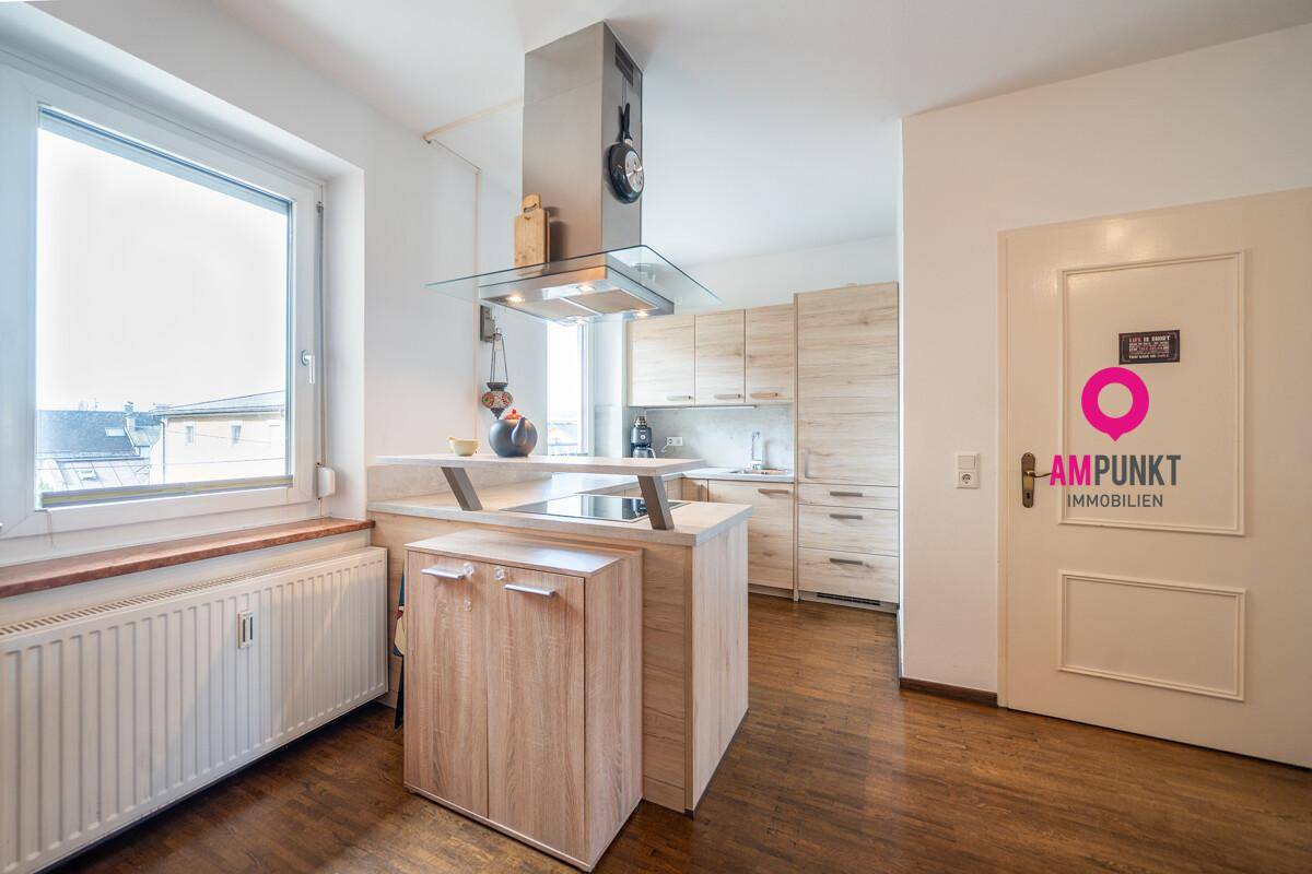 Moderne Küche mit hellen Holzfronten, Kochinsel und Dunstabzugshaube, gut beleuchtet durch ein großes Fenster.