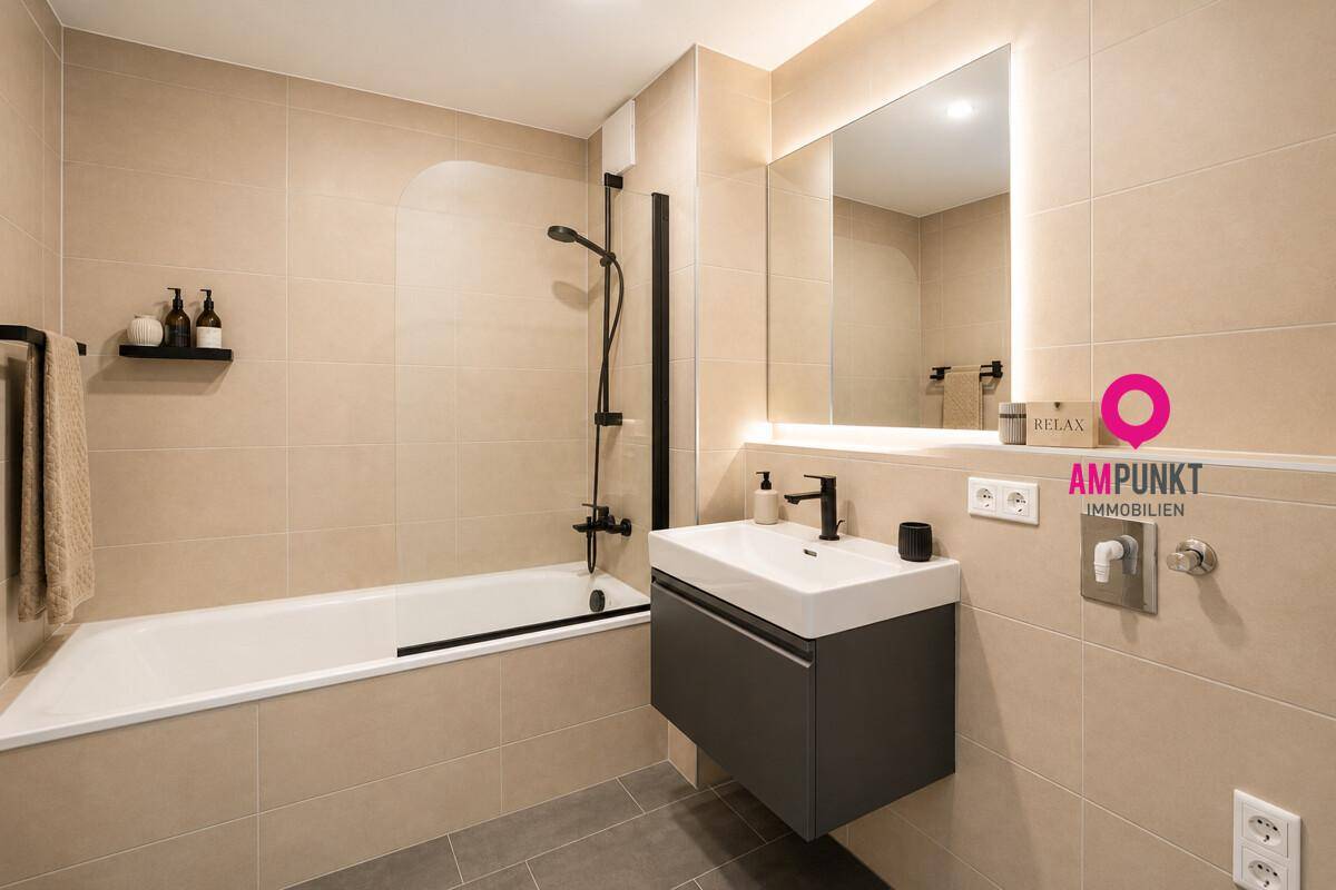 Modernes Badezimmer mit beigefarbenen Fliesen, Badewanne und beleuchtetem Spiegel.