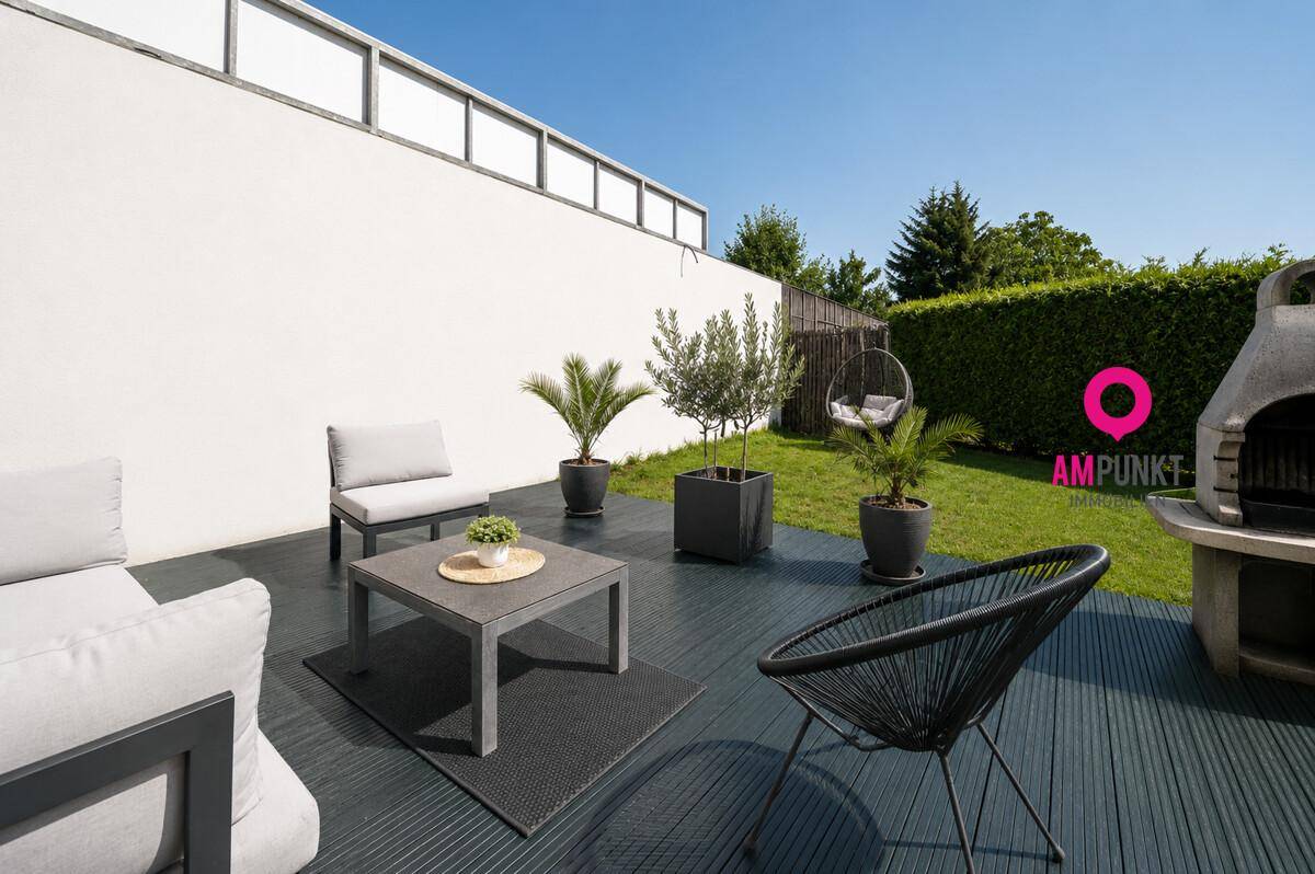 Sonniger Gartenbereich mit Terrasse, Grillplatz und Rasenfläche, umgeben von Hecken.