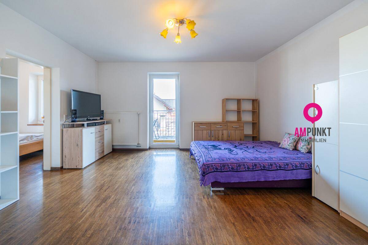 Geräumiger Wohnbereich mit Bett, TV-Möbeln und Zugang zu einem kleinen Balkon, hell und funktional eingerichtet.