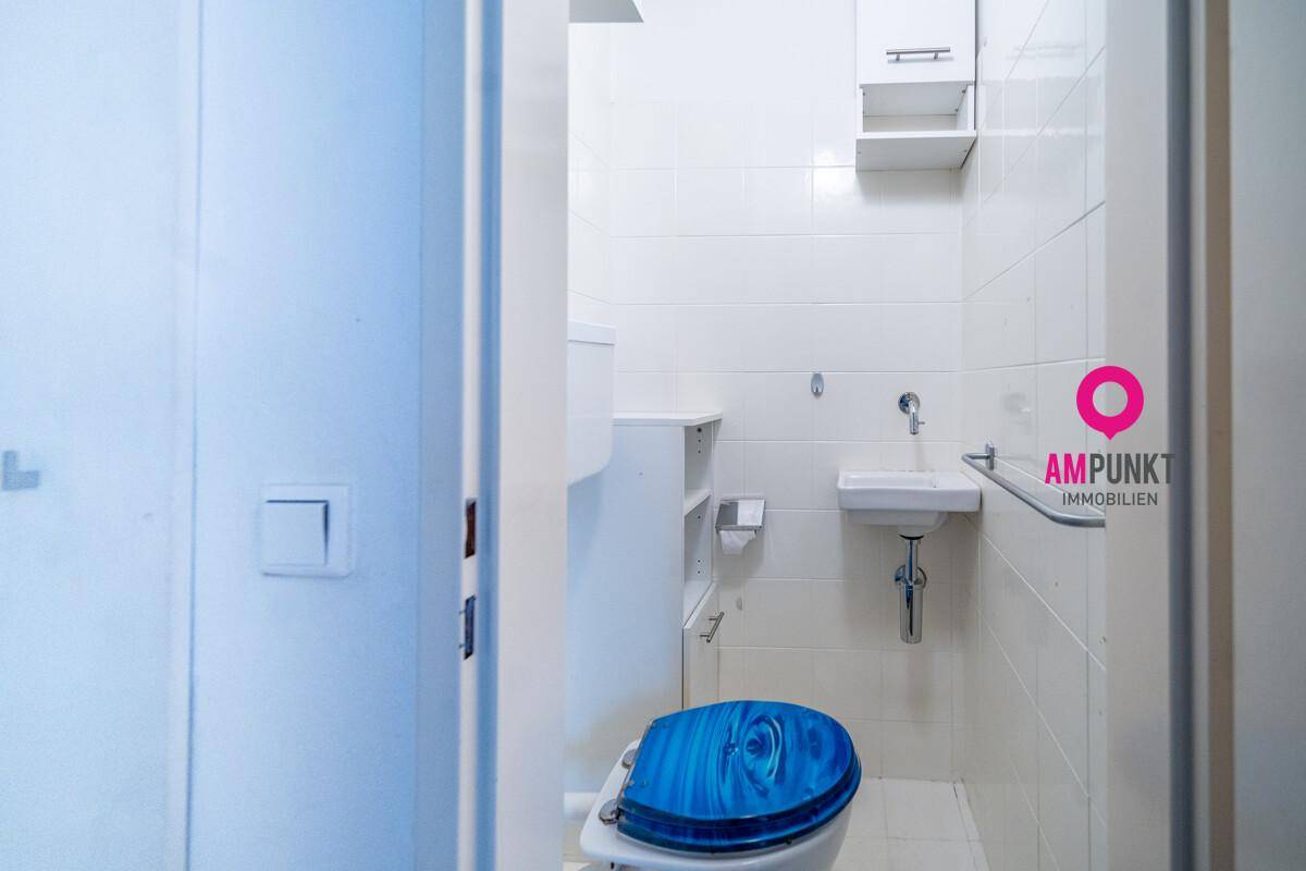 Separates WC mit Toilette und kleinem Handwaschbecken, vollständig gefliest in hellem Weiß.