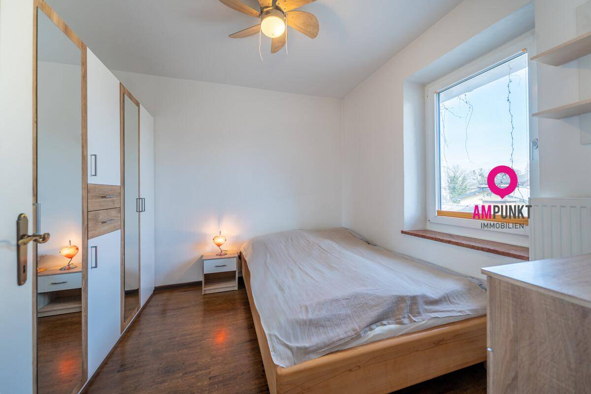 Schlafzimmer mit Holzbett, weißem Kleiderschrank mit Spiegeltüren und einem Fenster, ausgestattet mit Deckenventilator.