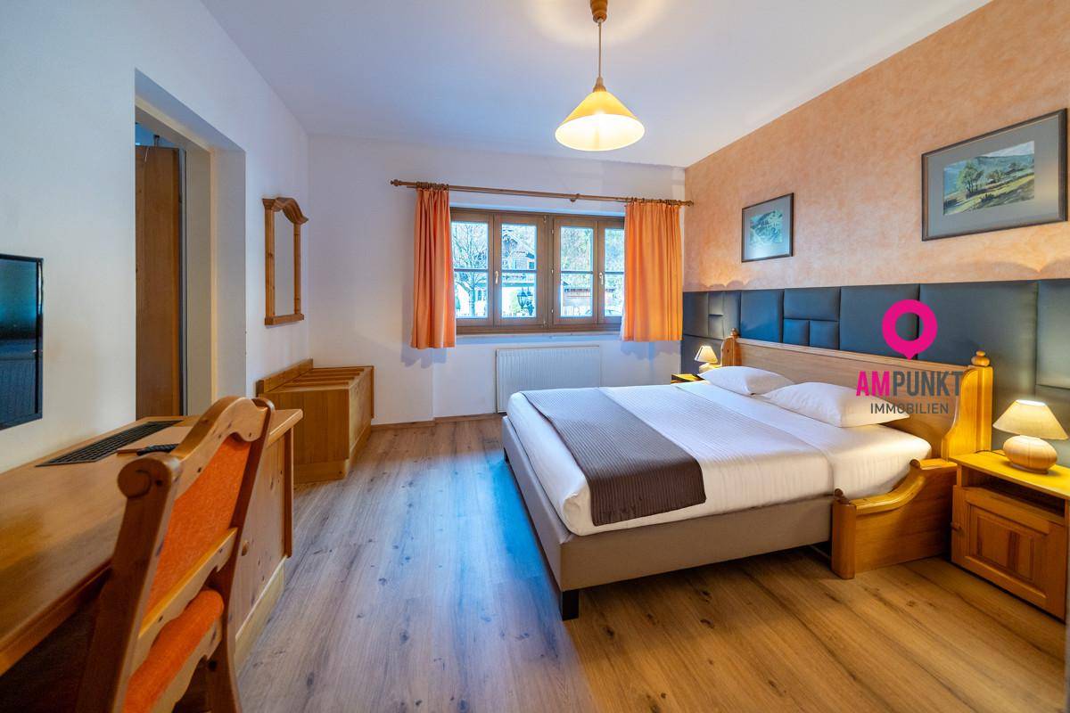Komfortables Schlafzimmer mit Holzboden, Doppelbett, Schreibtisch und orangefarbenen Vorhängen, mit Blick ins Grüne.