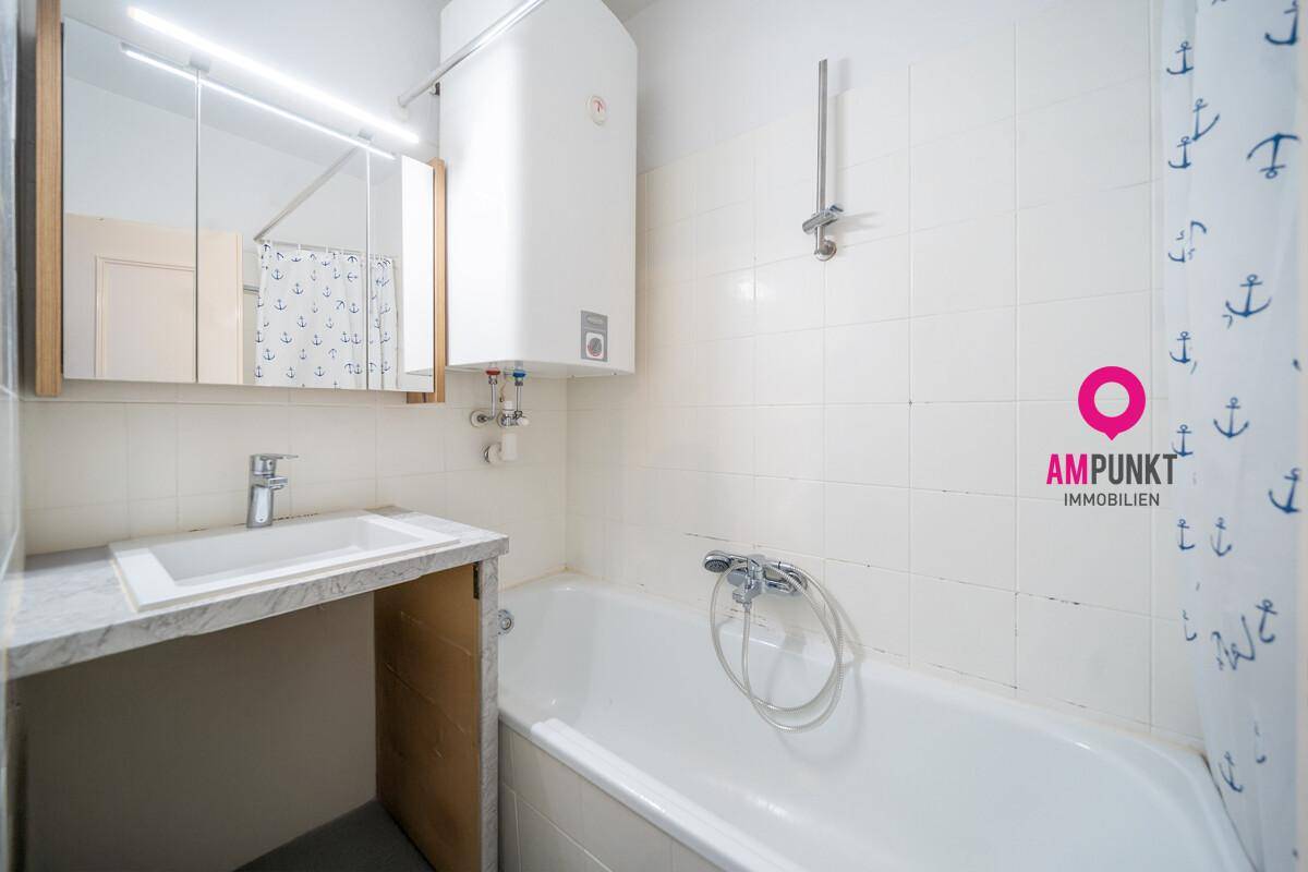 Badezimmer mit Badewanne, Waschbecken, Spiegelschrank und Warmwasserboiler, komplett gefliest in Weiß.