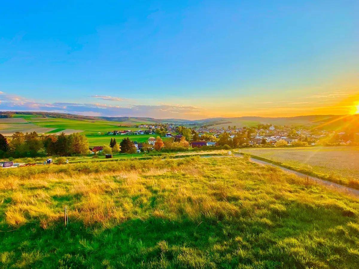 Panoramablick über grüne Felder und ein Dorf in der Ferne bei strahlendem Sonnenuntergang.