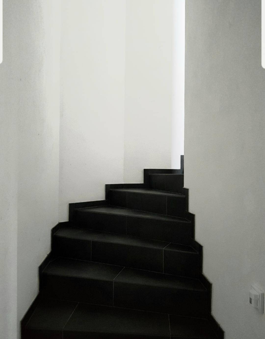 Treppenhaus mit dunklen Fliesenstufen und schlichten weißen Wänden, die nach oben führen.