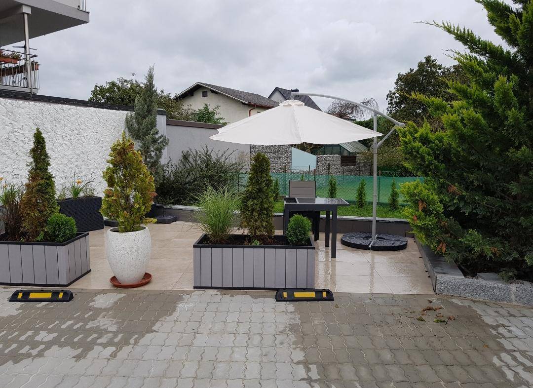 Gepflasterte Terrasse mit Gartenmöbeln, Sonnenschirm und dekorativen Pflanzen in modernen Töpfen.