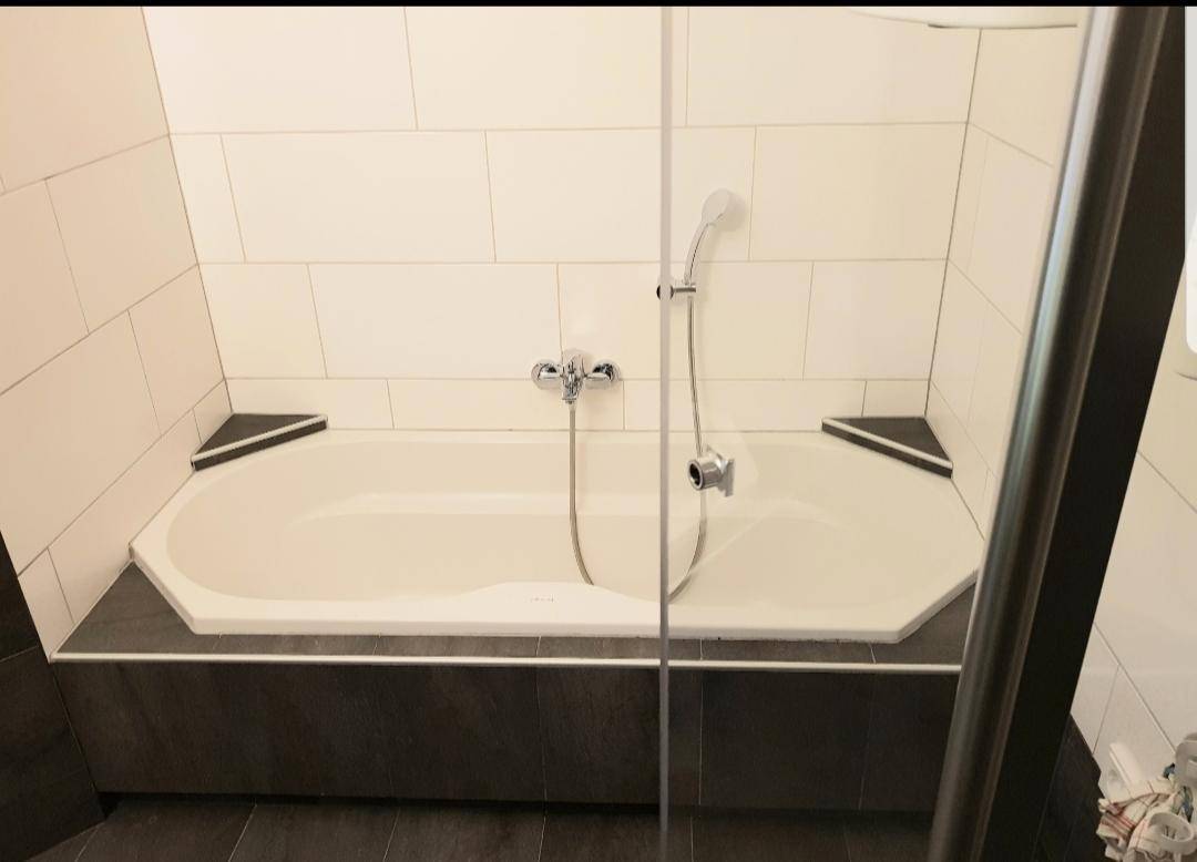 Helle Badewanne mit Duschfunktion, umgeben von weißen und grauen Fliesen.