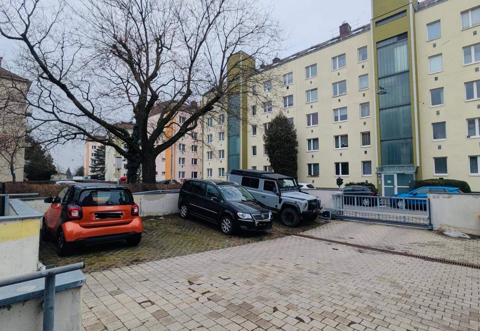 Blick auf einen Außenparkplatz mit geparkten Fahrzeugen und einer Wohnanlage im Hintergrund.