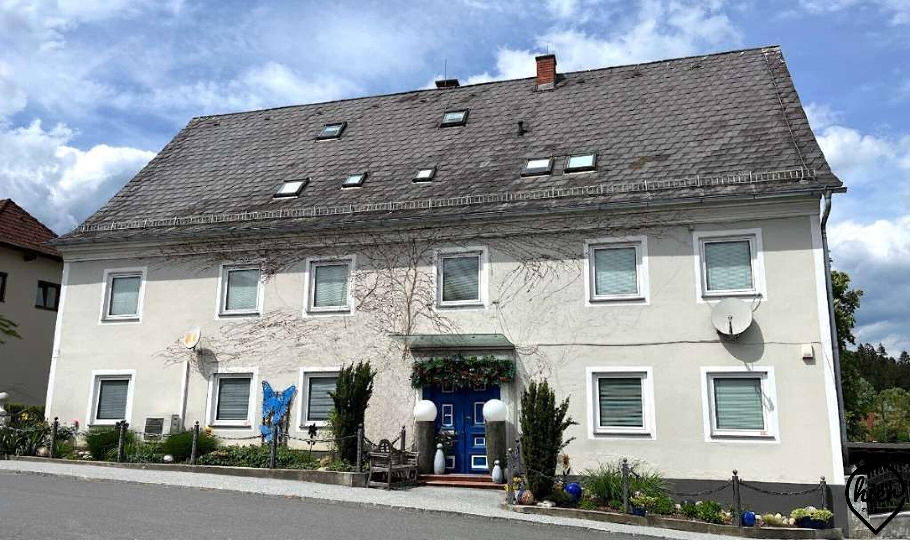 Historisches Herrenhaus mit Satteldach, vielen Fenstern und blauer Eingangstür, umgeben von Grün.