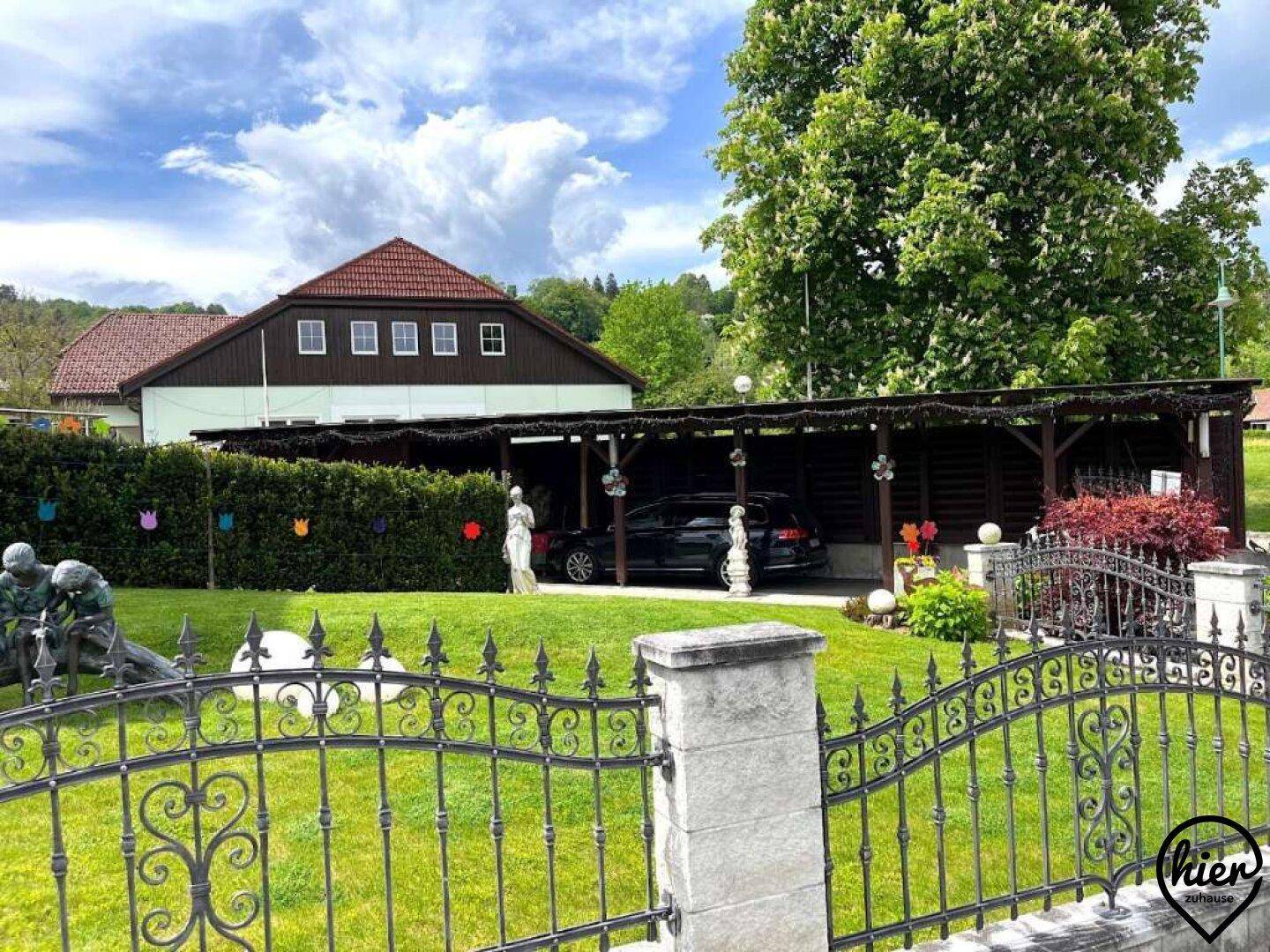Gepflegter Vorgarten mit Carport, schmiedeeisernem Zaun und Skulpturen unter blauem Himmel.
