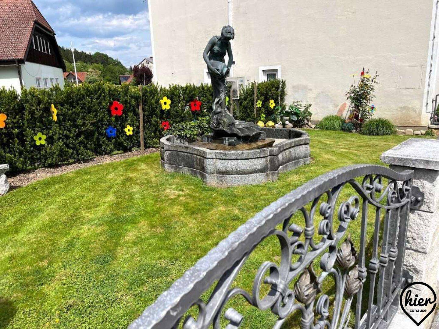Grüner Vorgarten mit einem dekorativen Brunnen und schmiedeeisernem Zaun vor dem Haus.