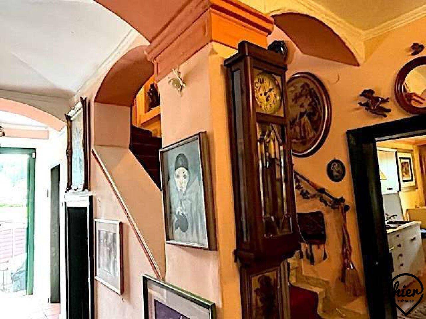 Traditioneller Flur mit Holztreppe, antiker Standuhr und zahlreichen Gemälden an den Wänden.