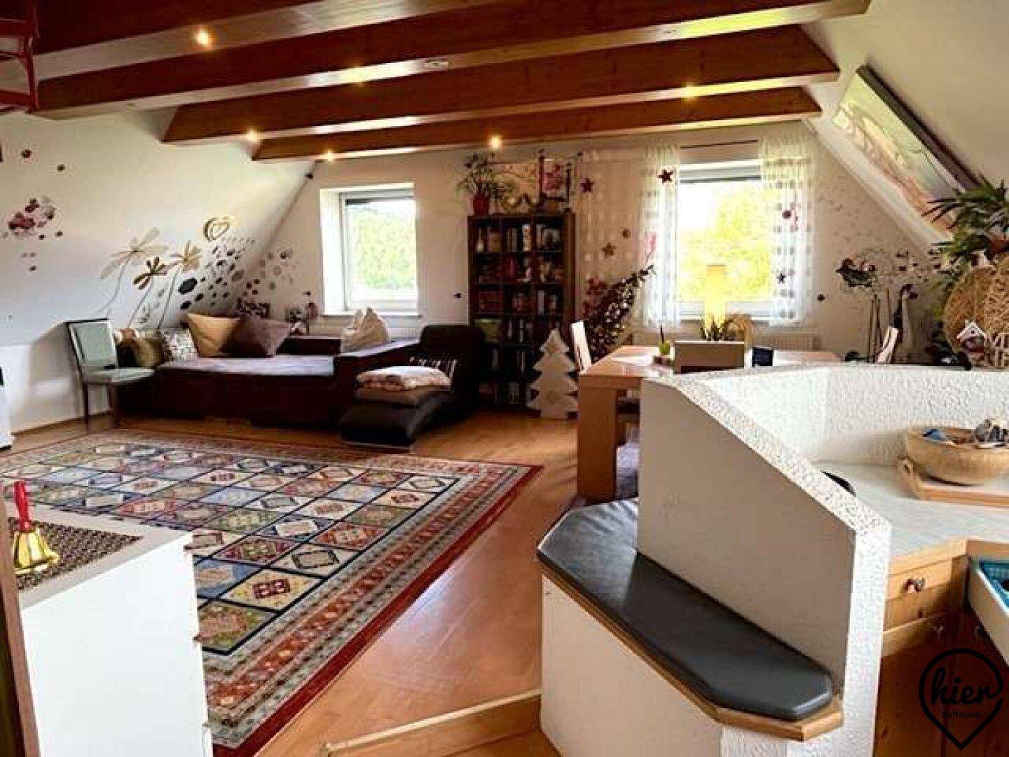 Gemütlicher Wohn-Essbereich im Dachgeschoss mit Holzbalken, Teppich und viel Tageslicht durch die Fenster.