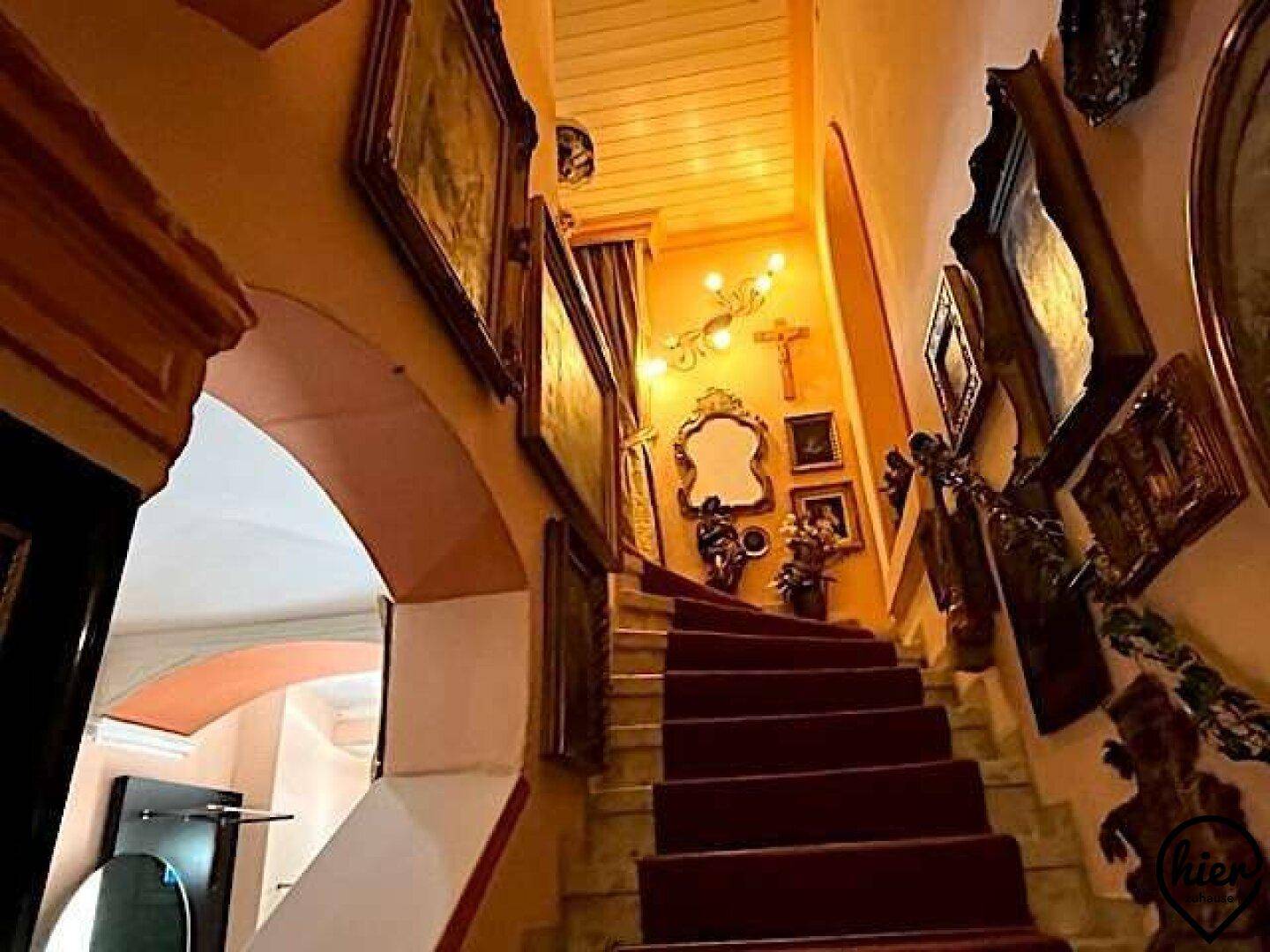 Elegante Treppe mit rotem Teppich und kunstvollen Gemälden, die zu den oberen Etagen führt.