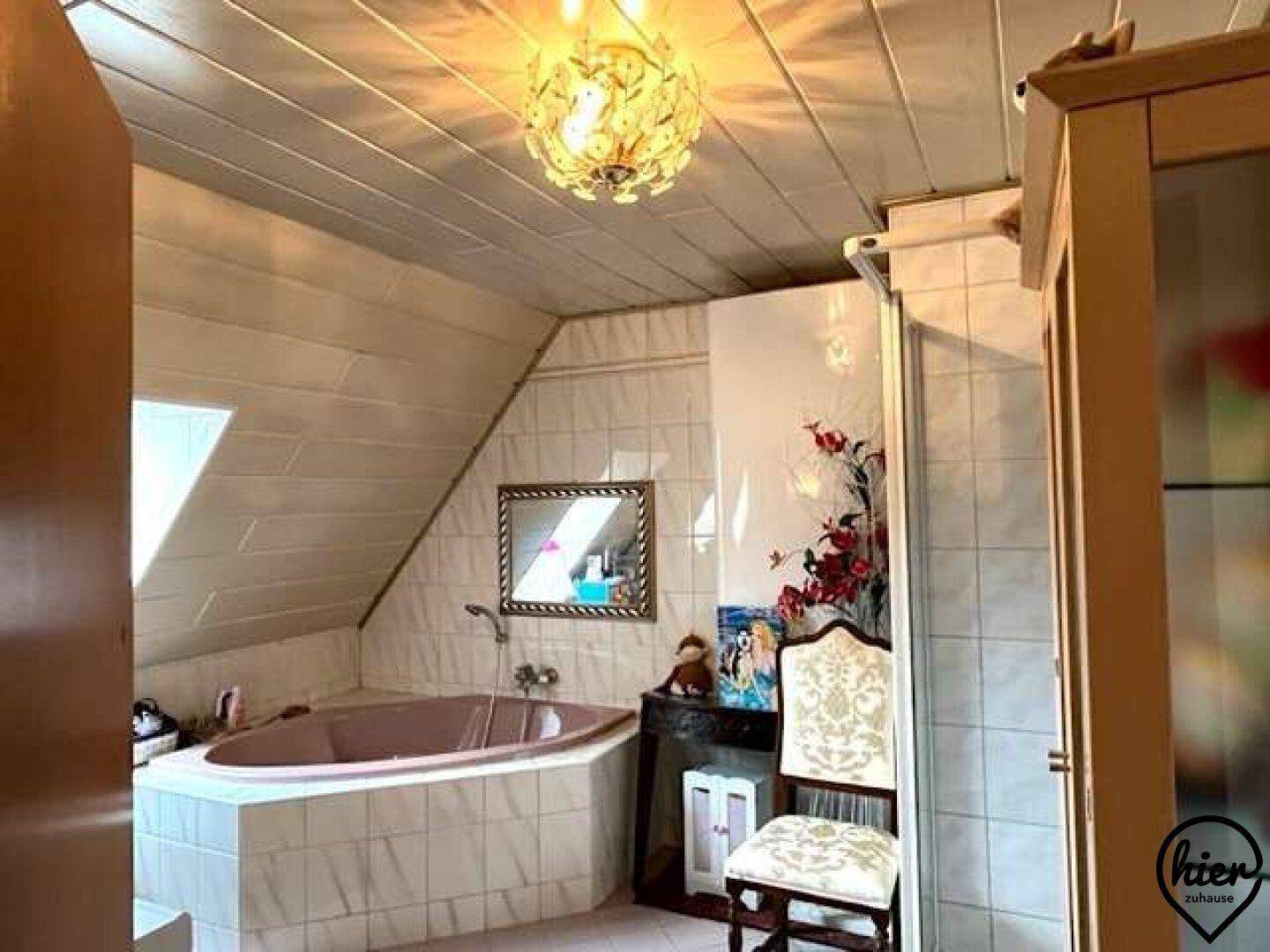Badezimmer im Dachgeschoss mit Eckbadewanne, hellen Fliesen und Holzdecke unter einem Dachfenster.