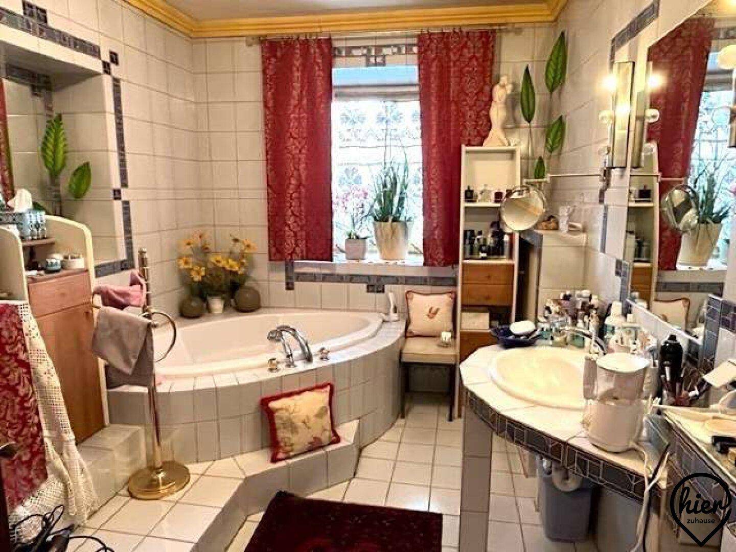 Geräumiges Badezimmer mit Eckbadewanne, Doppelwaschbecken, Fliesen und Fenster mit roten Vorhängen.