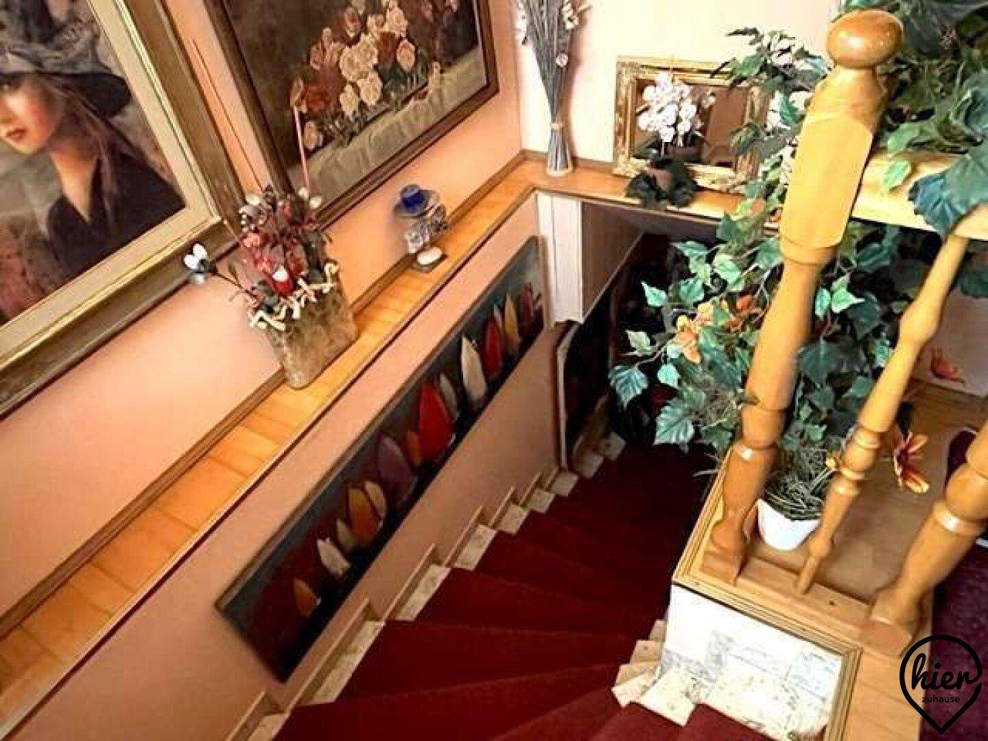 Treppenhaus mit rotem Teppich, Holzgeländer und dekorativen Gemälden entlang der Wand.